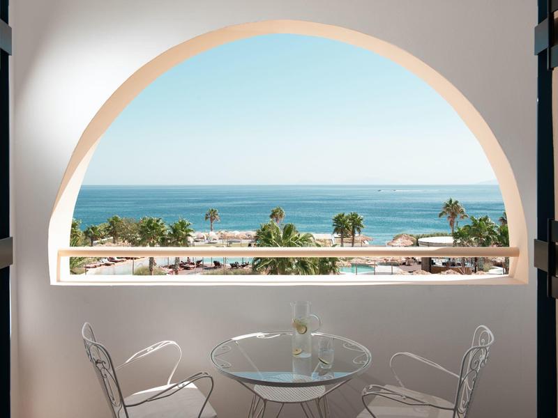 Výhled obloukovým oknem na palmy, pláž a klidné moře se stolem a židlemi v popředí.