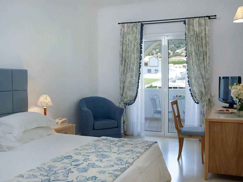 Světly hotelový pokoj s postelí, modrým křeslem, dřevěným stolem a balkonem s výhledem na bílé budovy.