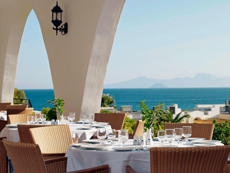 Venkovní terasa restaurace s výhledem na moře, bílé stoly a proutěné židle pod oblouky.