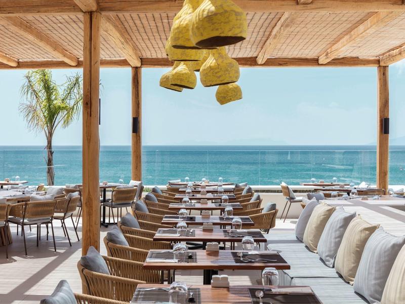 Přímořská restaurace s dřevěnými stoly, proutěnými židlemi a závěsnými lucernami pod dřevěnou střechou.