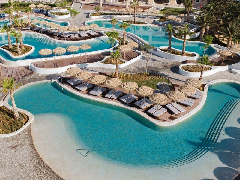 Velký hotelový bazén s několika částmi, lehátky a palmami obklopený písčitými plochami.
