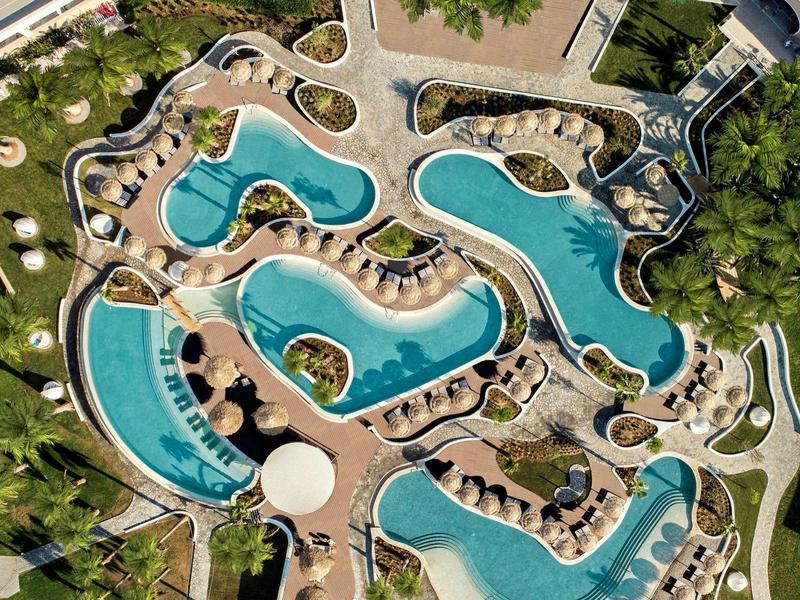 Přehled křivolaké hotelového bazénu s lehátky a zelení kolem.