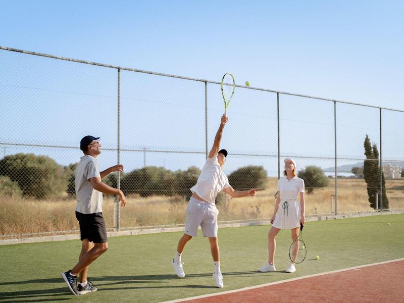 Tři lidé hrají tenis na venkovním hřišti za slunečného dne.