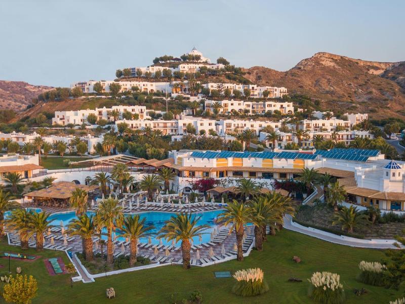 Hotelový resort s několika bazény a bílými budovami na kopci před horami při západu slunce.