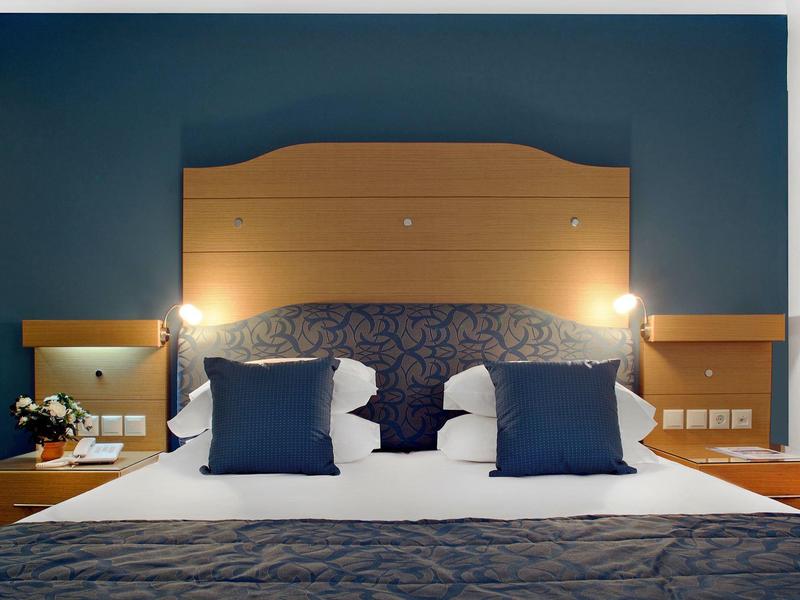 Symetrický hotelový pokoj s modrým čelem postele, dvěma lampami a ozdobnými polštáři na posteli.