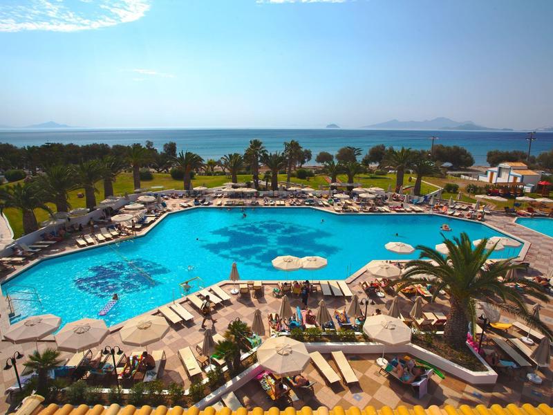 Velký hotelový bazén s lehátky, palmami a výhledem na moře za jasné oblohy.