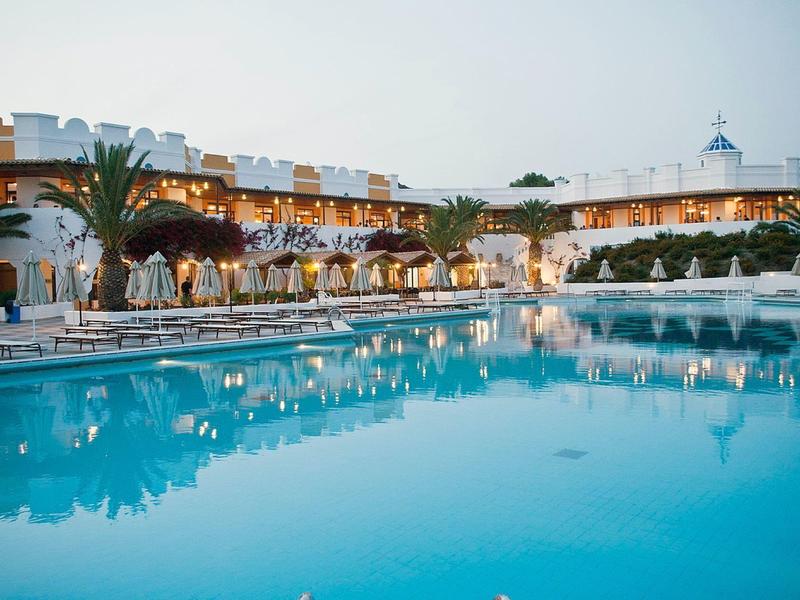 Hotelový bazén s lehátky a osvětlenou budovou v pozadí za soumraku.