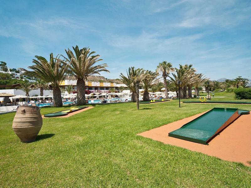 Zelený trávník s palmami a venkovním mini golfem před hotelem pod modrou oblohou.