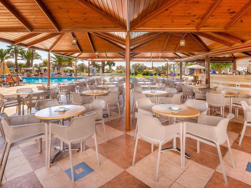 Venkovní restaurace s bílými stoly a židlemi pod dřevěnými střechami u bazénu.