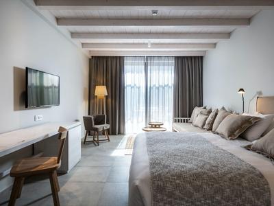 Licht ingericht hotelkamer met groot bed, bureau, stoel en balkon met gordijnen.