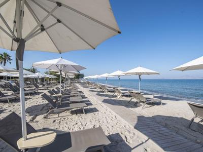 Reihe von Liegestühlen mit Sonnenschirmen am Sandstrand neben ruhigem Meer unter klarem Himmel.