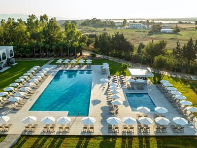 Modernes Hotel mit zwei Pools, weißen Liegestühlen und grüner Landschaft im Hintergrund.