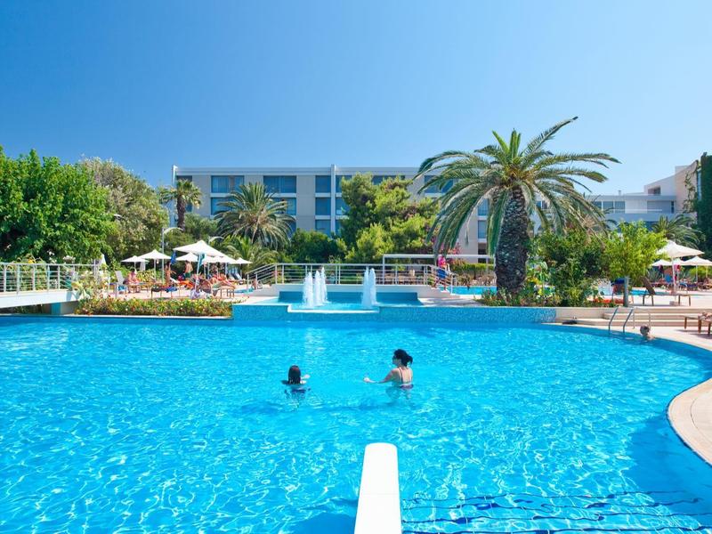 Groot hotelzwembad met fonteinen, palmbomen en zwemmers onder een heldere hemel.