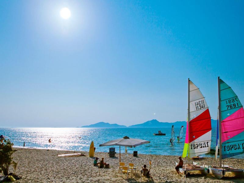 Zonnig strand met mensen die ontspannen en kleurrijke zeilboten bij het water.
