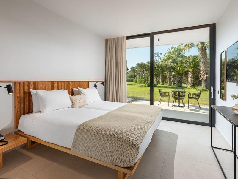 Moderne hotelkamer met groot bed, glazen schuifdeur naar groene tuin.