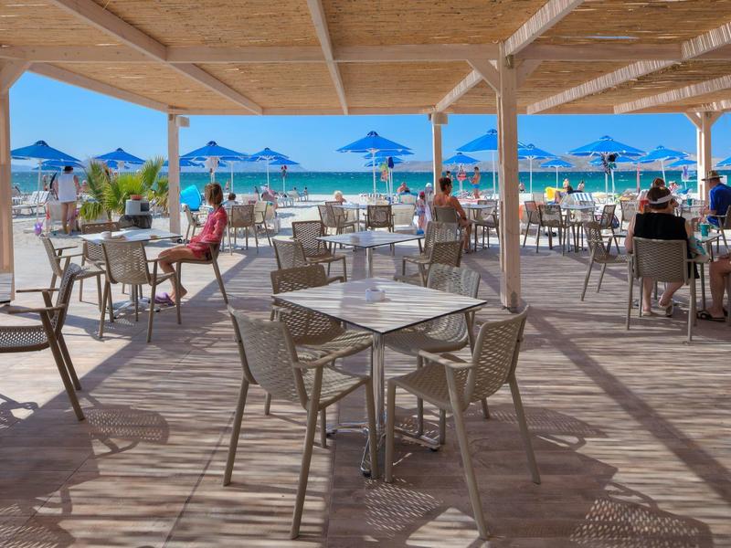 Buitenterras van hotel met tafels, stoelen in de schaduw, gasten genieten van strand en blauwe parasols.