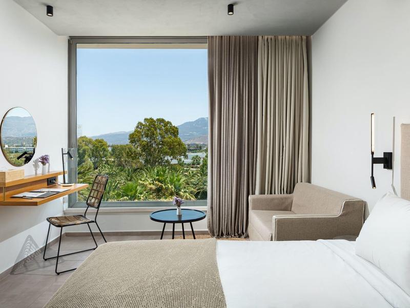 Moderne hotelkamer met groot raam, beige bank, bed, bureau, stoel en uitzicht op buiten.