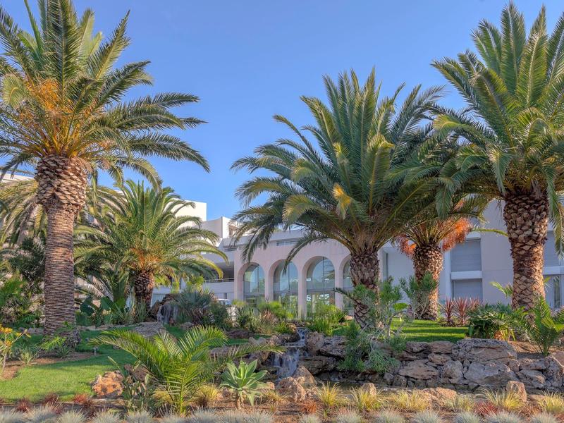 Hotel tuin met palmbomen en wit gebouw met bogen onder een blauwe lucht.