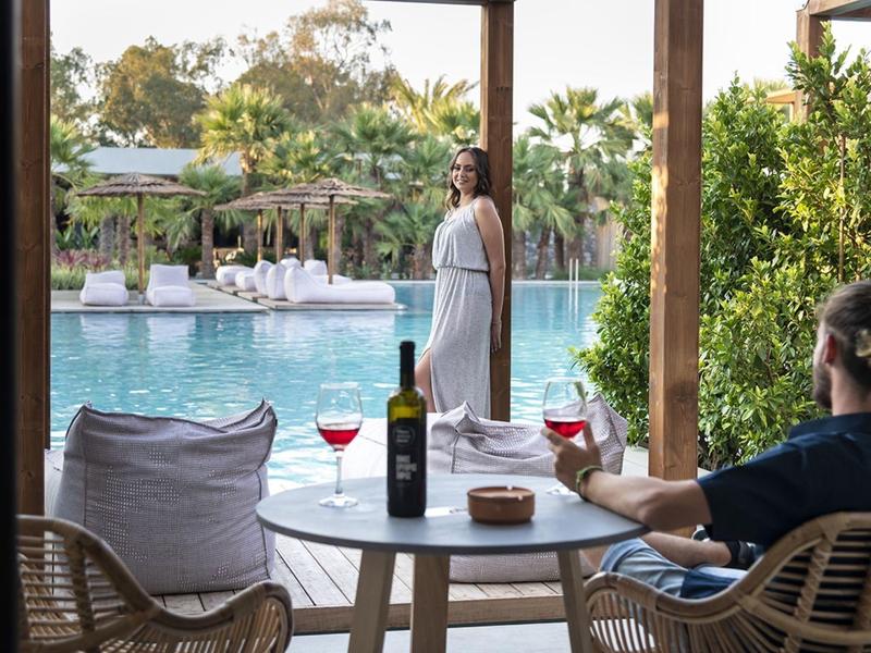 Man en vrouw genieten van drankjes aan een tafel met uitzicht op een zwembad en weelderig groen.