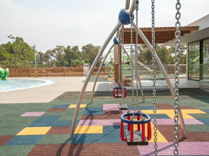 Lege kinderschommels op kleurrijke rubberen tegels bij een zwembad en gebouw.
