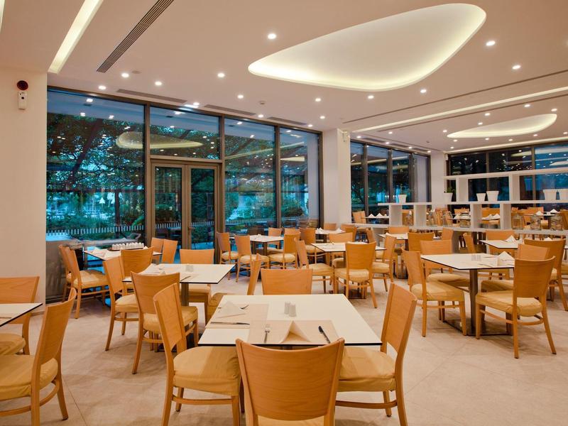 Ruime eetzaal van hotel met houten stoelen, tafels, grote ramen en moderne plafondlampen.