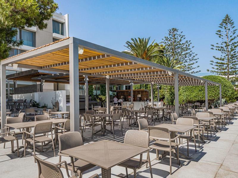 Buitenterras van hotel met tafels, stoelen en schaduwrijke pergola op zonnige dag.