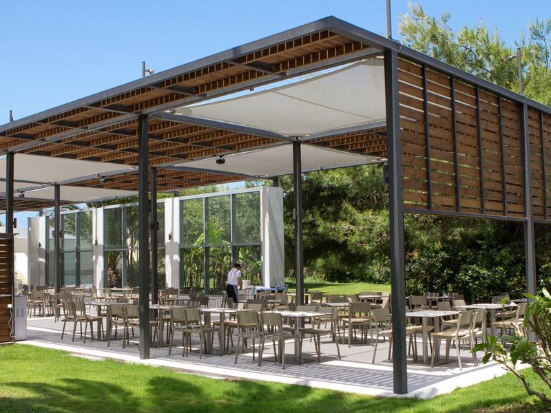 Buitenterras met tafels, stoelen en gedeeltelijk dak in een tuinomgeving.