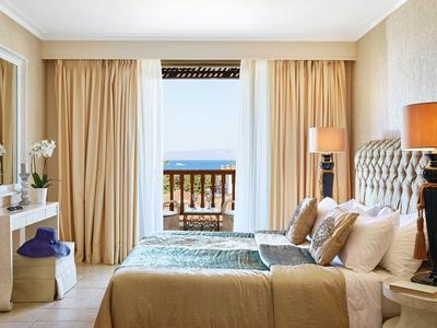 Bright hotel room with a large bed, beige curtains, and a balcony overlooking the sea.