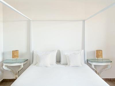 Minimalist white bedroom with canopy bed, two pillows, bedside tables, and wicker lamps.