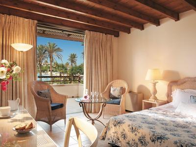Cozy hotel room with wooden beam ceiling, double bed, seating area, and palm tree view.