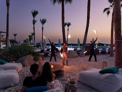 Menschen sitzen am Strand bei Sonnenuntergang neben einer Feuerstelle und Palmen.