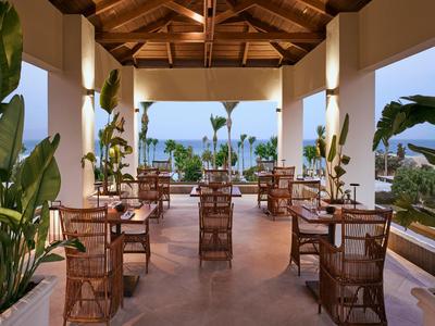 Open dining area with wooden chairs and sea view, surrounded by green plants.
