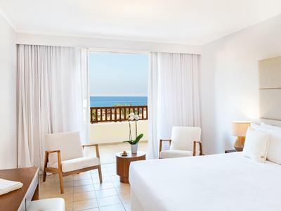 Bright hotel room with double bed, chairs, and sea view through large window.