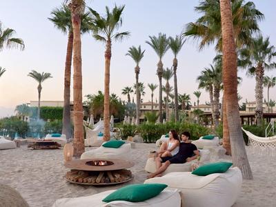 Couple relaxes on beach loungers with palm trees and hotel in background at sunset.
