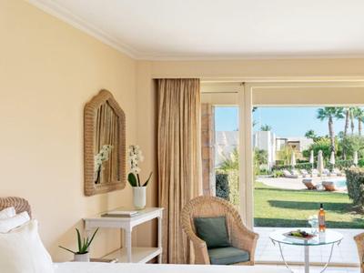 Elegantes Hotelzimmer mit weißem Bett, Sitzbereich und Blick auf einen Garten mit Pool.