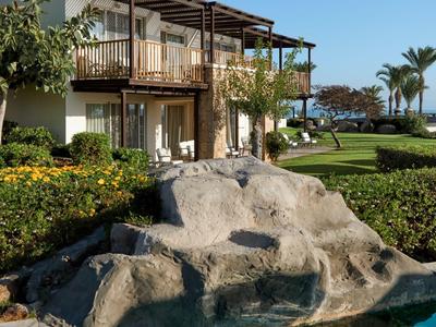 Hotelgebäude mit Balkon über einem Kieselsteinbecken und grünem Garten unter klarem Himmel.