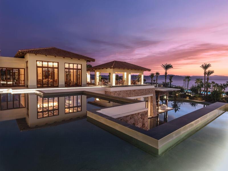 Modern hotel pool at sunset with reflective water and palm trees.