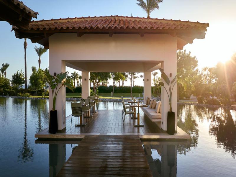 Covered seating area on wooden platform surrounded by water and palm trees in sunlight.