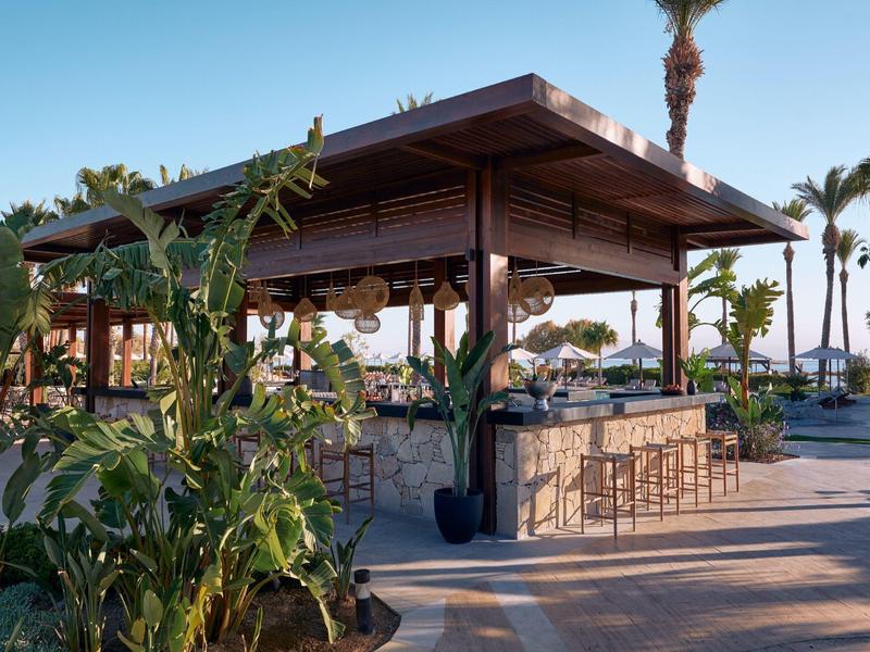 Open beach bar with wooden roof, stools, and counters, surrounded by green plants and palm trees.