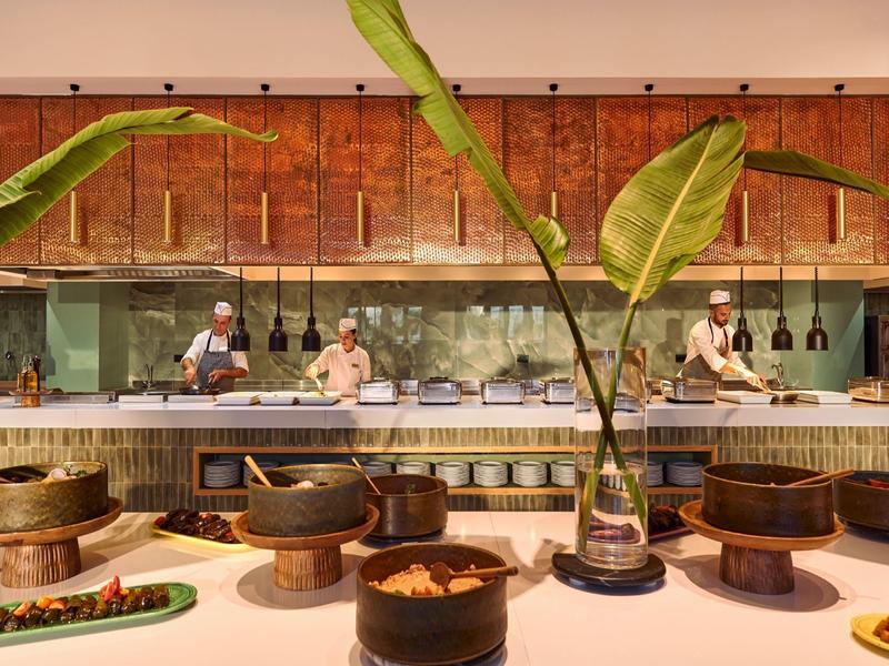 Modern hotel kitchen with chefs, wooden shelves, and plants in the foreground.
