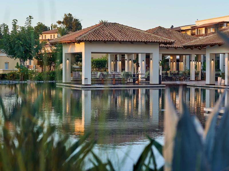 Hotel with pavilion and terrace by calm water reflecting buildings and plants in the foreground.