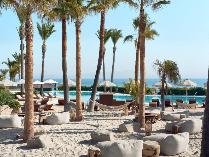 Beach resort with palm trees, bean bags, and parasols by the sea under clear skies.