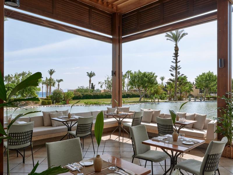 Outdoor restaurant seating area overlooking a pool and lush greenery in daylight.