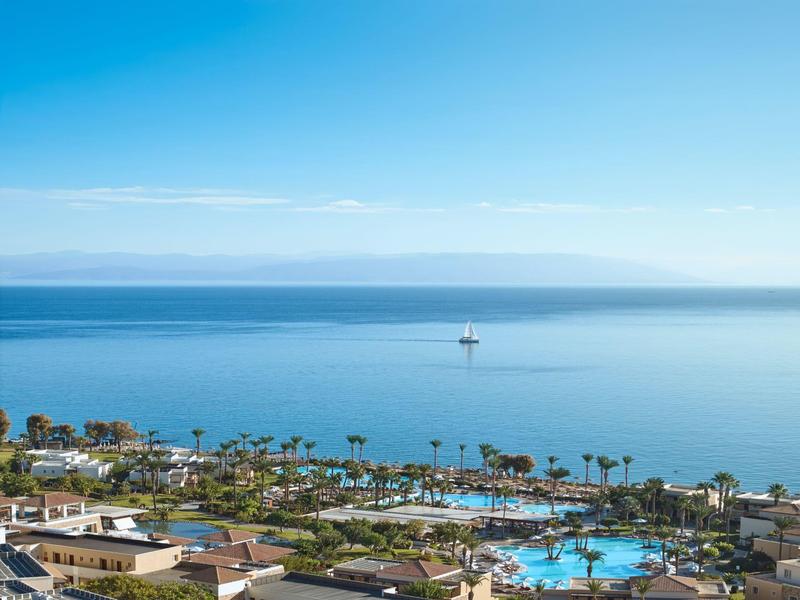 View of a resort with pool, palm trees, and calm sea under a clear blue sky.