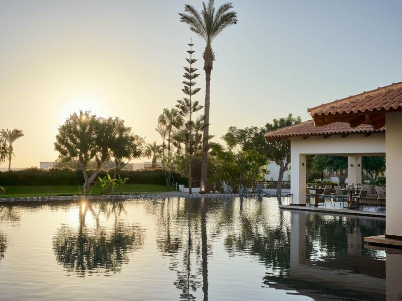 Sunset over a calm pool with palm trees and hotel lounge area reflected in water.