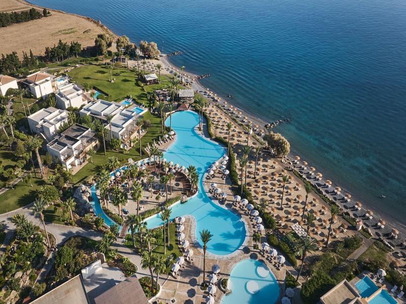 Aerial view of a resort with large pools, palm trees, and sea view.