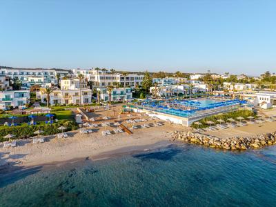 Modernes Hotel mit Pool an einem Sandstrand und klarem blauem Wasser bei sonnigem Himmel.