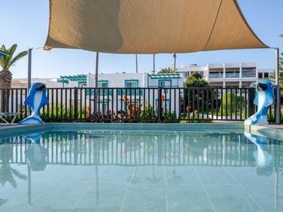 Poolbereich mit Schutzsegel und zwei blauen Delfinwasserfontänen vor einem Hotelgebäude.