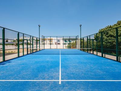 Blauer Paddle-Tennisplatz mit Netz, umgeben von einem grünen Zaun und Gebäuden im Hintergrund.