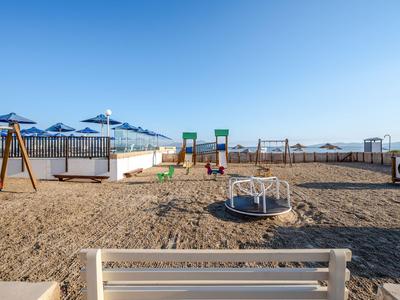 Strandspielplatz mit Sand, Spielgeräten und blauen Sonnenschirmen unter klarem Himmel.